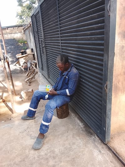 A man in blue overalls with white reflective stripes on the legs sits on a wooden stump next to a large, dark metal gate. He is holding a clear plastic container with a white lid, which appears to contain a light-colored liquid. A spoon is also visible in his hand, and he is looking down at the container. His hair is short and appears to be dusted with a light-colored substance. The man is wearing grey slip-on shoes. The ground is concrete and dirt. In the background, there is a concrete wall and some building materials. The scene appears to be outdoors, possibly a workshop or industrial area, given the setting cues. The lighting suggests it is daytime, with shadows indicating the sun is present. The overall mood is one of a break or a moment of rest. No visible text can be discerned.