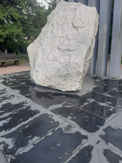 An imposing, roughly hewn, light-colored boulder is the central focus of this outdoor scene, positioned on a wet, dark-tiled plaza. The boulder exhibits a textured surface with visible cracks and variations in tone, suggesting it is natural stone. To the right of the boulder, a series of tall, slender, grey vertical pillars are present.
In the background, lush green trees with dense foliage dominate the upper left portion of the image, with hints of buildings and possibly other greenery visible through the leaves. A wooden park bench with slatted seating and dark legs sits on a reddish-brown paved area to the left of the boulder and trees.
The plaza beneath the boulder and extending forward is comprised of large, dark square tiles, many of which are wet and reflective, creating dark, irregular patches that contrast with the drier, lighter grey areas of the tiles. The overall impression is of a public space that may have recently experienced rain. The scene is devoid of any people or animals. The location is confirmed to be Shelekhov, Russia.