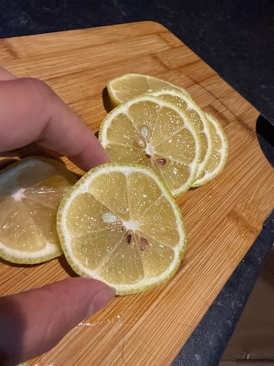 A high-angle, close-up shot shows a person's hand holding a lemon slice on a wooden cutting board. There are several other lemon slices on the board, arranged in a slightly overlapping manner. The lemons appear ripe, with a pale yellow flesh and a thin, light green rind. The hand is holding one of the slices, displaying it to the viewer. The cutting board has a distinct wood grain pattern with a light brown color. A dark surface, possibly a countertop, is visible in the background. There is no information to suggest the location is Great Yarmouth, United Kingdom.