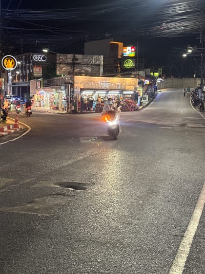 The image depicts a nighttime scene on a street in Karon, Thailand. The street is paved and shows signs of wear, including some potholes. On the left side of the street, there are shops with illuminated signs. One prominent sign reads "OZO." Further down the street, there are other establishments with colorful displays of merchandise, possibly souvenirs or clothing. A person wearing a light-colored patterned shirt and a face mask is riding a scooter, with its headlights on, moving from left to right. To the right of the scooter, other scooters are parked along the roadside. In the background, there are more buildings, streetlights, and a significant amount of overhead electrical wiring. On the right side of the street, a paved walkway leads uphill, with a few pedestrians visible. The overall atmosphere is that of a commercial street at night, likely a tourist area.