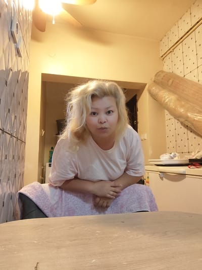 A woman with blonde, somewhat disheveled hair is sitting at a table in a room. She is wearing a light pink t-shirt and a pink and white patterned blanket is draped over her lap. Her hands are clasped together in front of her. The room appears to be a kitchen or living area, with tiled walls, a refrigerator visible in the background, and some rolled items wrapped in plastic leaning against the wall. A ceiling fan is visible in the upper left corner of the image. The lighting is warm and somewhat dim. The location is identified as Sarıyer, Turkey.