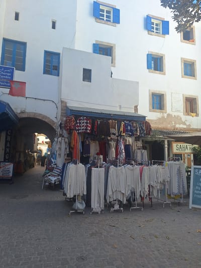 In Essaouira, Morocco, a street scene unfolds with traditional architecture and commerce. A white building with blue shutters frames the upper portion of the image, its facade punctuated by several windows of varying sizes. A Moroccan flag, red with a green star, is visible on the left.
Below the building, a bustling marketplace is in full swing. Clothing racks overflow with garments, including intricately patterned tops and long, flowing tunics. Some items are displayed on mannequins, while others hang from racks and from the overhang of a stall. A sign for "SAHA HE" is partially visible on the right, suggesting a shop or restaurant. The cobblestone street is partially covered in fallen leaves, indicating an outdoor setting. People are present in the scene, though their details are indistinct. The overall impression is one of a vibrant, lively marketplace within the historic medina of Essaouira.