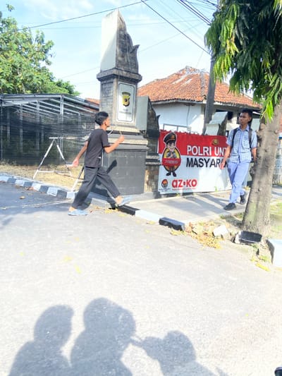 The image shows two young men walking on a paved street in front of a building with a sign that reads "POLRI UNT MAS YARA." The man on the left is wearing a black t-shirt, black pants, and black shoes. He is walking away from the camera. The man on the right is wearing a blue and white checkered shirt, blue pants, and black shoes. He is walking towards the camera. The sign is red and white with a cartoon image of a police officer in the top left corner. The sign is attached to a brick building with a stone column in front of it. There is a green tree on the right side of the image. The location of the image is in Purwakarta, Indonesia, based on the provided coordinates. The shadows of the two young men are clearly visible on the ground. The location is a street in Purwakarta, Indonesia. The environment is an urban setting.