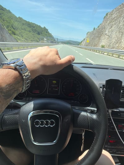 The image shows a driver's perspective from inside a car, with a hand on the steering wheel. The car is on a highway with a mountainous landscape visible on both sides. The driver is wearing a watch on their left wrist. The car's dashboard displays speed, temperature, and other information. The weather appears to be sunny. The location is identified as Bolesestra, Montenegro.