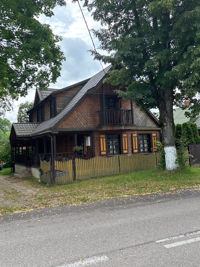 The image depicts a two-story wooden house with a dark gray roof, located in the village of Trześcianka, Poland. The house has a covered porch running along the front, supported by dark wooden posts. A light-colored wooden fence encloses the property in front of the house. The house features dark-trimmed windows, some with decorative shutters. A large tree stands to the right of the house, with its trunk painted white at the base. The foreground shows an asphalt road with some scattered leaves. The sky is overcast. No people are visible in the image.