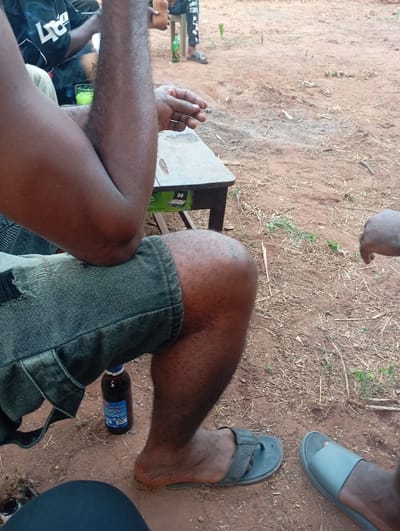 A group gathers outside, evident by the dirt ground. A man in shorts sits with an elbow on a small table, a cigarette held in his hand. A beer bottle sits on the ground next to his leg. Another man wearing flip flops stands in the background, and the feet of other individuals are partially visible. The scene implies a casual gathering with drinks and relaxation.