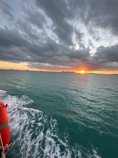 The image shows a view from a boat in the ocean near Thong Nian, Thailand. The sun is setting, casting a warm orange and yellow glow across the horizon and reflecting on the water. Dark, dramatic clouds fill the sky, creating a contrast with the vibrant sunset. In the foreground, the wake of the boat is visible, with white foam churning in the turquoise water. A portion of an orange life preserver is visible on the left side of the frame. Distant islands can be seen silhouetted against the sunset.
