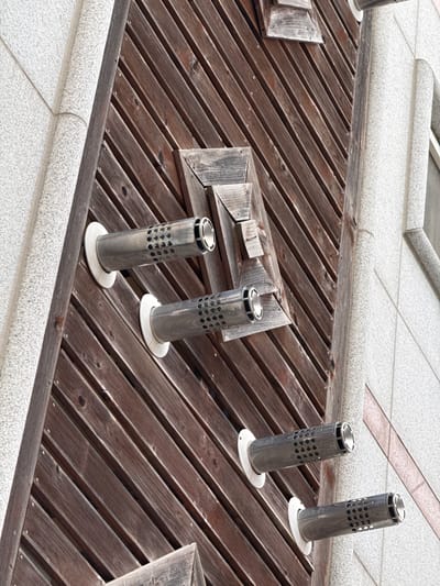 The image shows a close-up, high-angle view of a textured, light gray building facade adjacent to a section of dark brown wooden planks. Three cylindrical metal vents with perforated ends protrude from the wooden surface. These vents are angled downwards, with circular white flanges at their bases where they attach to the wood. The wooden planks are arranged diagonally, creating a slatted effect. The building facade features a speckled, grainy texture. No people or actions are visible. The scene appears to be an exterior architectural detail. This specific location is the exterior wall of the Mokpo Modern History Museum 1 in Mokpo-si, South Korea.