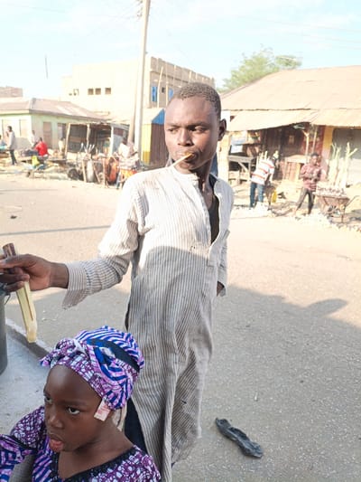 A man stands on a street, sucking on a sugar cane stick. He wears a long-sleeved, vertically striped shirt and holds another sugar cane piece. A young girl, wearing a patterned headwrap and shirt, stands beside him, looking to the side. The street scene includes small shops and people in the background, with scattered items on the ground. Buildings are visible in the distance under a clear sky.