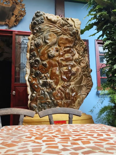 In Xuân Thành, Vietnam, a large, ornate, carved stone sculpture is displayed outdoors. The sculpture, primarily in shades of brown and beige, features a complex, irregular surface with what appear to be carved figures or natural formations. It stands on a golden pedestal. In front of the sculpture, a mosaic-tiled table with a terracotta and white pattern is visible, with two wooden chairs positioned behind it. The background consists of a bright blue wall, a red-framed window, and lush green foliage. To the left, a dark red door with glass panels and a circular silver doorknob is partially open. Above the door, a carved wooden decorative piece is affixed to the blue wall. The scene is set in what appears to be a courtyard or outdoor seating area.