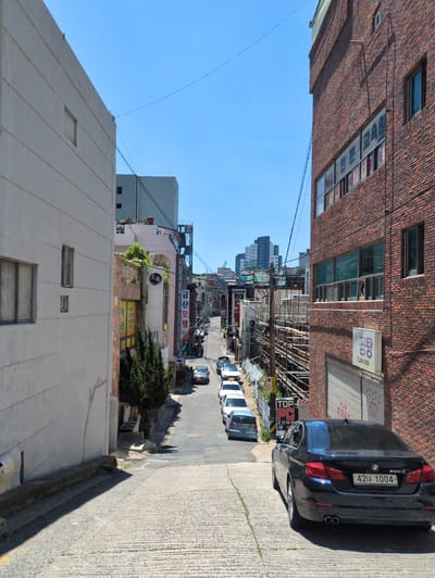 A street scene in Ulsan, South Korea. A narrow street is lined with buildings on either side. The buildings are constructed from brick and concrete. Utility wires are visible overhead. Cars are parked along the side of the street. Signs with Korean writing are visible on the buildings. A black BMW with a license plate "42너 1004" is parked. The sky is clear and blue.