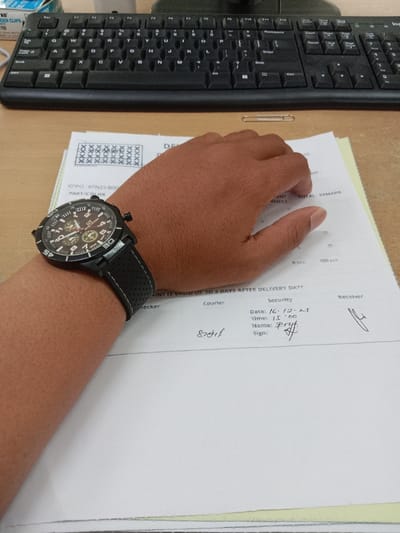 A black keyboard and a hand wearing a black watch with a brown face are positioned over a stack of papers on a desk. The top paper is a form with boxes filled with "X"s, and sections labeled "PART/COLOR", "REJECT", and "REMARK." Filled out security fields show the date as "16.12.25" and the time as "1.500". The name "Pryf" is printed, and a signature is scrawled beside it. The word "Receiver" is also present, with another signature next to it. A paperclip sits near the top of the page.