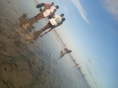 The image depicts a coastal scene in Shanzu ward, Kenya, featuring multiple individuals wading in shallow, clear water under a blue sky. In the foreground, three males stand partially submerged, with water reaching approximately their lower calves. The leftmost male wears an orange shirt and dark shorts, holding a dark, possibly deflated, tire inner tube. The central male wears a white basketball jersey with "LAKERS" and the number "3". The rightmost male wears a white and green basketball jersey with "RODGERS" and the number "12". Their reflections are visible on the water's surface, which contains sparse submerged seagrass. Further in the midground, another individual is seen partially submerged, also holding a dark tire inner tube. In the distant background, a horizontal line of numerous people extends across the water, indicating a populated shallow beach or tidal flat environment. The sky is predominantly clear blue with a few thin white clouds. The lighting suggests daytime.
