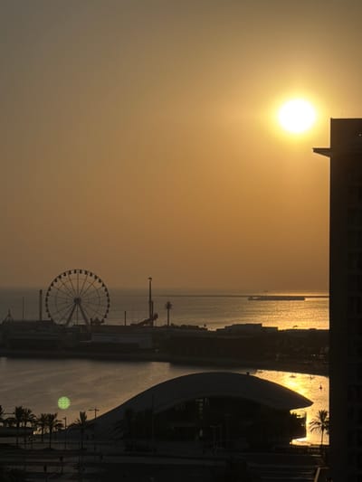 The image captures a sunset scene in Lusail, Qatar, with the sun prominently visible in the upper right quadrant of the sky. The golden hues of the sunset cast a warm glow over the entire scene. In the mid-ground, a large Ferris wheel stands silhouetted against the sky, suggesting an amusement area. The waterfront is visible, with calm water reflecting the warm light of the setting sun. A modern architectural structure with a curved roof is in the foreground, and palm trees are sparsely scattered along the waterfront. To the right of the image, a tall building is partially visible, also silhouetted against the bright sky. The overall atmosphere is serene and picturesque, typical of a coastal city at dusk.