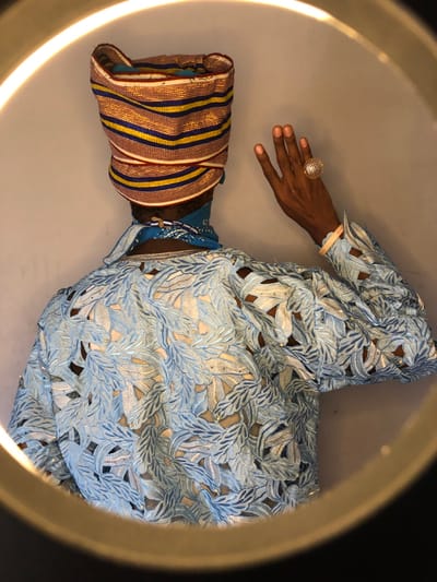 The image, captured in Ngleshi Amanfro, Ghana, displays the posterior view of an individual positioned against a plain, light-colored wall, partially encircled by the bright arc of a ring light. The person wears a structured headwrap composed of tightly folded fabric with alternating horizontal stripes in gold, blue, and red. A blue bandana is visible around the neck, beneath which a blue beaded necklace rests. The upper torso and right arm are clad in a light blue garment featuring an intricate pattern of cut-out leaf shapes, some edged with lighter blue or silver embroidery and adorned with small, reflective embellishments. Through these cut-outs, a warm-toned underlayer or skin is visible. The individual's right hand is raised, palm facing outward, with fingers slightly parted. A large, circular, gold-toned ring with a textured, mesh-like surface is prominent on the ring finger of the right hand. A pale, flat wristband is also present on the right wrist.