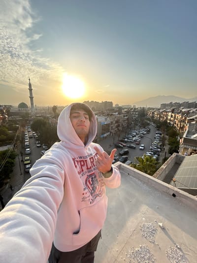 A male individual is depicted in a selfie, positioned on a concrete rooftop. He is wearing a light-colored hooded sweatshirt with a partially visible graphic design featuring "WESTERN EXPLORER" text and an American flag motif, along with dark trousers. His right hand is raised to chest height, with the middle finger extended. His gaze is directed towards the camera, with a slight smirk. The rooftop surface is light grey, showing scattered small white debris. To the immediate right, solar panels are visible on an adjacent lower structure, and a single palm tree is present.

The background reveals a densely built urban environment under a clear sky with scattered high-altitude clouds. The sun, a bright circular disc, is positioned in the upper-left quadrant, creating significant glare and lens flare, indicating either sunrise or sunset. A tall, slender minaret with a green dome is prominently visible in the upper-left background, identifying a mosque. Multiple streets wind through the city below, populated with numerous parked vehicles. Distant hazy mountains are observed along the horizon to the right. This scene is located within the city of Sarouja Municipality, Syria.