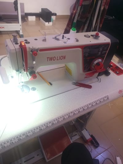The image shows a close-up of a white and red "Two Lion" brand industrial sewing machine placed on a table. The sewing machine is the primary object in focus, with its various components clearly visible, including the needle, presser foot, handwheel, and control panel. To the left of the sewing machine, a white industrial sewing machine stand is partially visible.

Several accessories are scattered around the sewing machine and on the table. A yellow pencil lies next to the sewing machine's needle, and a pair of small red scissors is positioned on the table. A spool of white thread is visible on the top of the sewing machine. A measuring tape, marked with inches and centimeters, hangs in the background, draped over a checkered fabric.

The table itself has a ruler-like marking along its edge, starting from inch 13 and extending beyond inch 32, indicating precise measurements. A red foot pedal is visible under the table. The setting appears to be an indoor workshop or studio, with tiled flooring visible in the background. The lighting suggests it is daytime, with a bright light source illuminating the sewing machine. The overall impression is one of a functional workspace, ready for sewing tasks. No people are visible in the image.