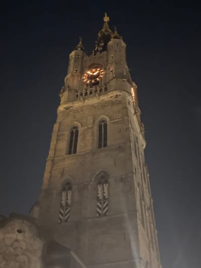 A stone tower with a clock face and gothic architectural elements stands tall against a dark night sky. The clock face, illuminated from within, shows the time as approximately 2:45. The tower has several windows and decorative features, and part of a stone structure, possibly a lower section of a building, is visible in the bottom left corner. The scene is illuminated by artificial lights, creating a warm glow on the tower. The image is likely of the Belfry of Ghent, a medieval bell tower located in the city of Ghent, Belgium.