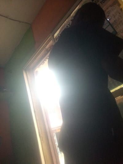 A full-body shot from a low angle captures a person silhouetted against a bright light source, possibly a doorway or window, in Shangisha, Nigeria. The person, wearing dark clothing, stands in profile to the right. The background is dominated by the intense light, with some indistinct shapes visible beyond it. To the left of the figure, a wall painted in shades of green and orange suggests an interior space. The overall lighting is high contrast, with deep shadows and blown-out highlights, making precise details difficult to discern.
