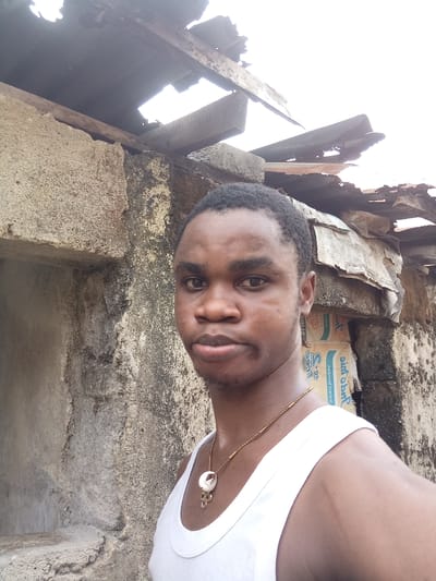 A medium shot depicts a young man in a white tank top with a gold chain necklace against a dilapidated structure. The background features a partially collapsed roof with visible wooden beams and corrugated metal sheets. Adjacent to the roof, a weathered concrete wall displays signs of age and neglect. Behind the man's right shoulder are faded boxes with reversed inscriptions. The setting appears to be in an area undergoing disrepair, potentially reflecting socio-economic conditions typical of some parts of Owerri, Nigeria. However, specific location in the city isn't readily determined. The man's direct gaze and the overall scene composition suggest a deliberate and intimate interaction with the viewer, potentially aimed at eliciting a response.
