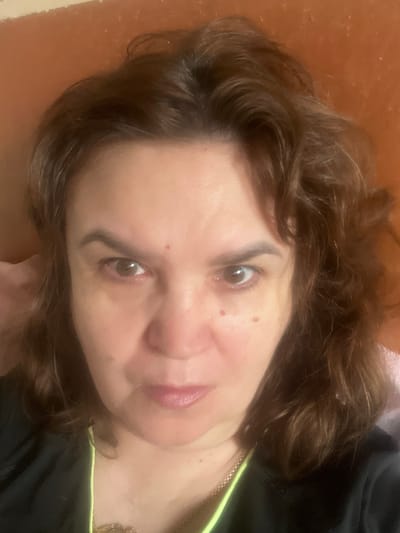 A medium close-up selfie shows a woman with brown, wavy hair, looking directly at the camera with a neutral expression. She is wearing a black shirt with a bright yellow neckline. A thin, gold chain necklace is visible. The background is out of focus, showing a textured, reddish-brown surface that could be a wall or furniture. There is no indication of the city of Tula, Russia, in the image.