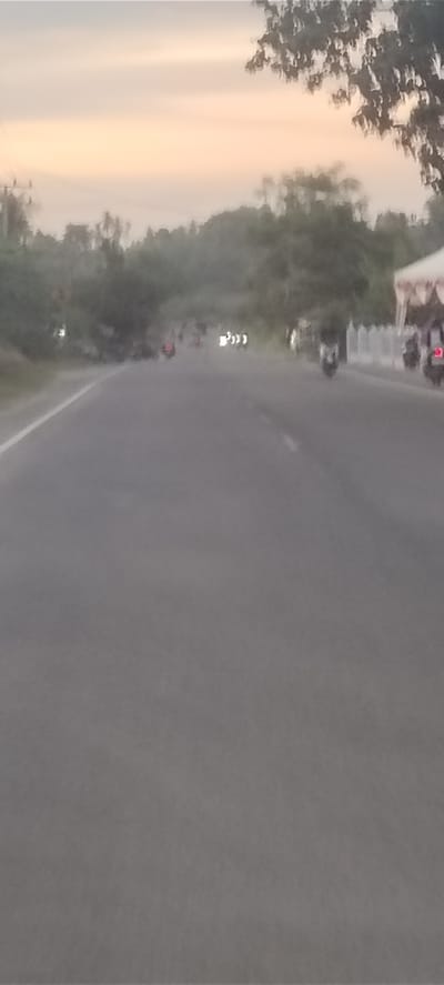 The image shows a road in Seuneubok, Indonesia. Several motorbikes are on the road. There are trees on either side of the road. The sky is a pale orange color, suggesting it is either sunrise or sunset. A white structure, possibly a tent, is visible on the right side of the road.