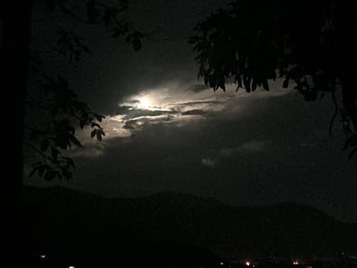 This is a nighttime photograph taken in the Ko Pha-ngan District of Thailand. The scene depicts a dark, mountainous landscape silhouetted against a sky illuminated by a bright full moon. The moon is partially obscured by wispy clouds, creating a dramatic and somewhat ethereal effect. The foreground is dominated by the dark shapes of tree branches and leaves, which frame the moonlit sky and add depth to the image. The leaves are silhouetted, indicating they are closer to the viewer than the moon. Below the mountains, faint lights suggest a settlement or town in the distance, hinting at human presence. The overall mood is serene and mysterious, evoking the quietude of a tropical night. The time of day is clearly night, and the presence of clouds suggests it might be humid or possibly a precursor to rain, though no rain is actively visible. The location is identified as Ko Pha-ngan District, Thailand, a place known for its natural beauty and nightlife. There is no visible text in the image.