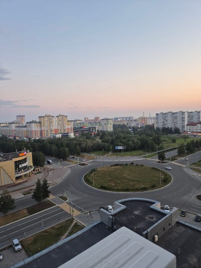 This image is an aerial view of a cityscape in Nizhnevartovsk, Russia, during twilight. The foreground features the rooftop of a building with a grey, textured surface and a parapet. Adjacent to this rooftop is a modern building with a grey facade and several cars parked on its roof, including a white SUV, a white sedan, and two dark sedans. A ladder is visible on the side of this building.
In the midground, a multi-lane roundabout dominates the scene, with cars circulating. Several crosswalks are marked with yellow and white stripes. A small, grassy island is in the center of the roundabout. Trees line the sides of the roads leading into and out of the roundabout.
Further back, a commercial building with a beige facade and the "DNS" logo is visible, along with signage in Russian. Beyond this, numerous multi-story apartment buildings in various colors and architectural styles stretch across the horizon. A dense forest separates the apartment buildings from the roundabout and commercial area. The sky is a soft gradient of pale blue and pink, indicating the early morning or late evening. The overall impression is of a residential and commercial district bathed in the soft light of dusk or dawn.
