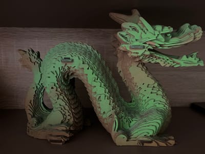 This media file features a layered, laser-cut cardboard model of a dragon. The dragon is predominantly a light tan color, with subtle green lighting that gives it an ethereal glow. It is coiled in a serpentine shape, with its head raised and mouth slightly open, appearing to be in a resting or alert pose. The construction of the model emphasizes layers and contours, creating a textured and dimensional effect. There are small, rectangular plates attached to the dragon's body, some of which have faint markings or text. One plate on the neck appears to have some embossed text, and near the base of the tail, the numbers "69" are clearly visible. The dragon is placed on a dark, flat surface, possibly a shelf or table. Behind the dragon, a textured, horizontal surface suggests a wall or backdrop, also dimly lit. The overall lighting is low, creating a moody and somewhat mysterious atmosphere. The scene appears to be indoors, with no visible indication of the time of day or weather conditions. There are no people visible in the media file. The specific location context of Kharkiv, Ukraine is not visually discernible from the image itself.
