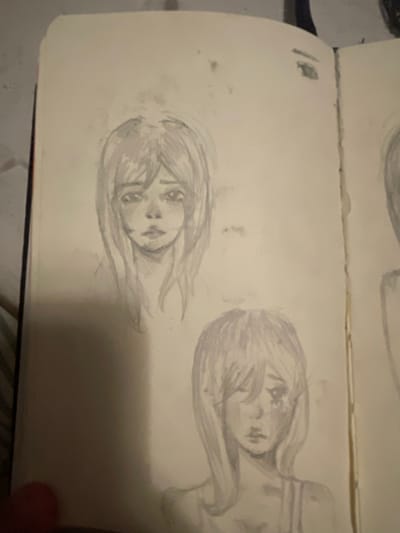 The image shows two pencil sketches of anime-style female faces in a sketchbook. The sketchbook is open, revealing two pages. The first sketch, on the left page, is a portrait of a young woman with shoulder-length hair and a melancholic expression. Her eyes are wide and downturned, and her mouth is slightly ajar. The second sketch, on the right page, is a similar portrait, but the woman's head is tilted, and one eye is partially covered by her hair, giving her a more mysterious or sad appearance. The sketches are rendered with soft shading, giving them a delicate and ethereal quality. The background of the sketchbook pages is plain white, with some smudges and faint lines that suggest the artist's process. There is a black binder clip visible at the top of the right page, holding the sketchbook shut. The image itself is a photograph, and the lighting suggests it was taken indoors. The context of Мухавецкі сельскі Савет, Belarus is not represented in the image.