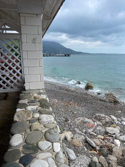 This is a photograph of a stony beach and the sea. In the foreground, a white lattice fence and a low wall made of large, rounded stones form a barrier. Beyond the wall, a pebble beach slopes down to the water. The sea is a muted blue-green, with small waves lapping at the shore and breaking around a few larger rocks. In the distance, a dark, tree-covered mountain rises steeply from the coastline. A pier or jetty extends into the water from the far shore. The sky is overcast with heavy, grey clouds, suggesting a cool or possibly rainy day. The overall mood is somewhat somber and tranquil. There are no people visible in the image. The lighting is diffused, indicating it is likely daytime, perhaps late afternoon or early morning, with the sun obscured by clouds. The location is identified as Abaata, Abkhazia.