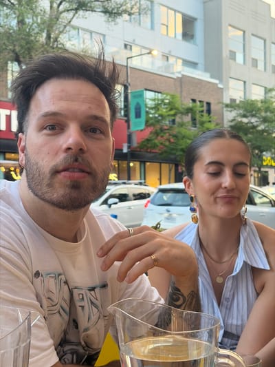 In this medium shot, a man and a woman are seated at an outdoor table, likely at a restaurant on a street in Montreal, Canada, given the style of the buildings and foliage. The man, in the foreground and to the left, is looking directly at the camera. He has short, dark, messy hair, a beard, and is wearing a white t-shirt with a graphic design on it. He has two rings on his right hand, one on the index finger and one on the ring finger, and a tattoo on his left forearm. The woman, to the right, is looking down and to her left, with her eyes closed, and a slight smile. She has her dark hair pulled back and is wearing a light blue and white striped halter top. She has large, multi-colored earrings and is wearing a necklace with a circular pendant. In front of them are two glass carafes of water, one of which contains lemon slices. Behind them, streetlights and trees are visible, along with the facades of buildings and cars, suggesting an urban setting. The overall impression is a candid moment captured during a meal or drink outdoors.