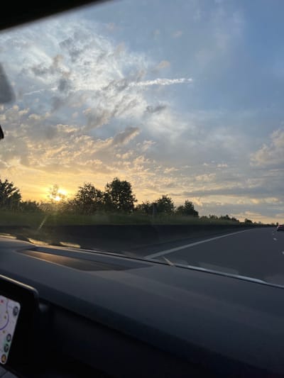 This image captures a highway scene during either sunrise or sunset, viewed from inside a vehicle, located in what is identified as Schifflange, Luxembourg.
The upper two-thirds of the frame are dominated by a sky displaying a mixture of clear blue patches and numerous clouds. The clouds nearer the horizon are illuminated by a strong, golden-orange light emanating from a partially obscured sun. This light source, located on the left side of the image, casts a significant glare. Clouds in the upper sky appear more diffuse and scattered, exhibiting lighter grays and whites.
The middle ground features a multi-lane highway, with at least two lanes visible in the direction of travel, delineated by solid and dashed white lines. A dark, low barrier or median separates the highway from the adjacent landscape on the left. Beyond this barrier, a dense line of deciduous trees and green foliage runs parallel to the road. The bright sun is visible through and behind these trees. On the far right, in the distance, a red passenger vehicle is discernible on the highway.
The foreground is occupied by the interior of the vehicle from which the photograph was taken. The dashboard, appearing dark gray or black, extends across the lower portion of the image, showing various contours and surfaces. Reflections or glare from the windshield are present in the upper left corner of the dashboard area. A portion of a digital display, likely a navigation system showing map-like graphics and icons, is visible in the bottom left corner, though its details are unreadable. No individuals are visible in the frame.