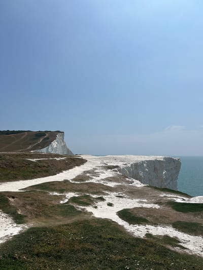The image depicts a coastal landscape under a clear blue sky. In the foreground, a grassy area with patches of white chalky soil is visible. A dirt path winds through this area, leading towards the cliffs. The middle ground is dominated by a dramatic white chalk cliff face that plunges into the turquoise sea. Further in the background, another, larger chalk cliff stretches along the coastline, with paths visible on its grassy summit. The sea is calm with small ripples. The scene appears to be set at Seaford Head, a prominent landmark in Seaford, United Kingdom, known for its white chalk cliffs and coastal paths. No people or animals are explicitly visible in the frame. The lighting suggests it is a sunny day.