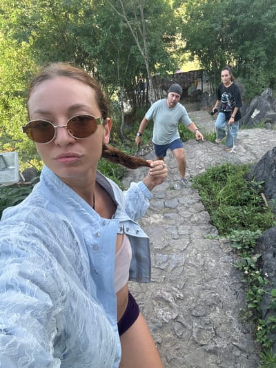 The image, taken at a high angle, shows a selfie of a person wearing round sunglasses and a light blue lace jacket. The person is holding a lock of hair. In the background, there are two other individuals on a stone path: one wearing a black t-shirt and jeans, and another in a light green t-shirt and blue shorts, pointing. The surrounding environment includes trees, stone structures, and vegetation. The location is in Đồng Văn Commune, Vietnam.