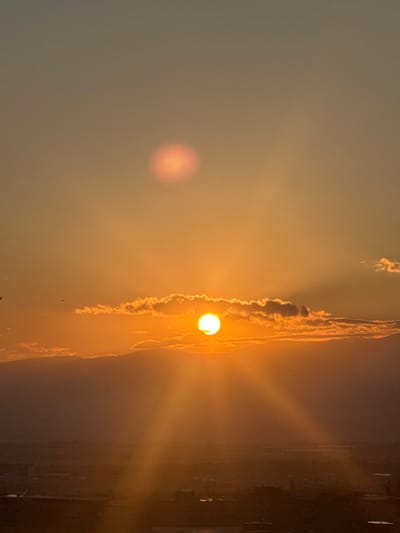 The image captures a sunset over the city of Abovyan, Armenia. The sun, a bright orange orb, is partially obscured by a line of clouds near the horizon. Sunbeams radiate outwards from the sun, creating a dramatic effect against the hazy orange sky. A faint, circular lens flare is visible higher in the sky. In the foreground, the silhouette of the city is visible, with buildings and infrastructure dimly lit by the fading sunlight. Several birds are scattered across the sky. The overall impression is one of a serene and beautiful natural phenomenon occurring over an urban landscape.