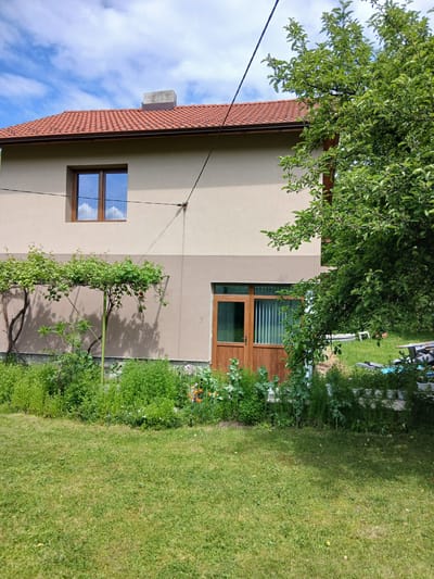 A two-story building in Ribaritsa, Bulgaria, features a tan facade, a red-tiled roof, a window on the lower level, and a wooden door on the upper level. A tree obscures part of the upper facade. A smaller tree is partially espaliered against the building. Green lawn is present with flowers and other greenery. Power lines are visible.