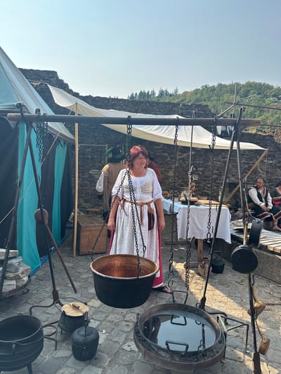 The image depicts a historical reenactment event likely taking place in Vianden, Luxembourg, given the prominent castle in the background. In the foreground, a woman in a white medieval-style dress with a brown belt and pouch stands near a large, hanging copper cauldron. To her left, a tent with teal and black fabric is visible. In the mid-ground, another individual in a brown tunic and a green feathered hat is seen, along with a wooden chest. A table covered with a white cloth displays various pottery and utensils. Further in the background, a man in a dark vest and white shirt is seated on a wooden bench, and other figures are present, some in period attire. The ground is paved with cobblestones. The scene is set outdoors with a clear sky and a backdrop of stone walls and trees, suggesting a historical site or fortification.