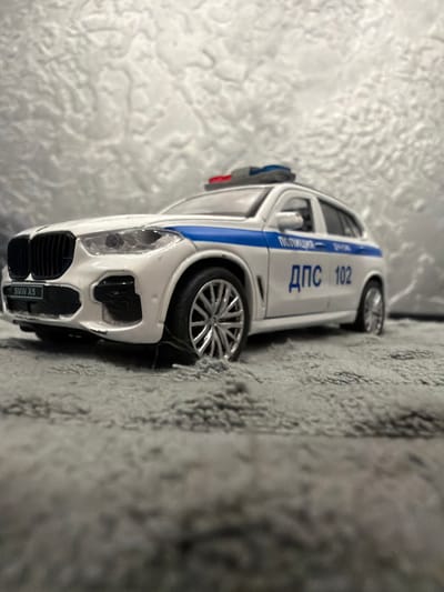 A white BMW X5 police car toy is positioned on a gray, fuzzy surface, likely a carpet or rug. The toy car is facing right, with its front right wheel slightly turned. It has a blue stripe running along its side, and Russian text which reads "ПОЛИЦИЯ" (Police) and "ДПС 102" (Road Patrol Service 102). The car has a visible license plate with "BMW X5" written on it. A red and blue emergency light bar is mounted on the roof. The background is a textured, light-colored surface. There is no indication of Mytishchi, Russia, or any specific urban environment in the image.