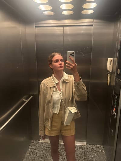 A young woman stands in an elevator, taking a mirror selfie. She is wearing a beige jacket over a textured white top and mustard yellow shorts. A small white crossbody bag with the word "CELINE" is slung over her shoulder. The elevator walls are made of brushed metal, and the floor is a speckled grey tile. The lighting in the elevator comes from several circular overhead fixtures. The elevator is in a building in Manavgat, Turkey.