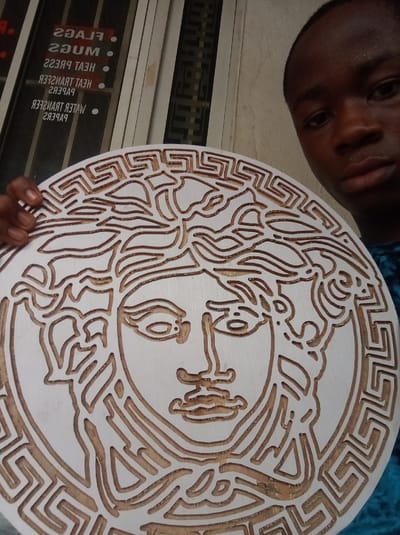 A young Black man is holding a circular, white, carved wooden plaque featuring the Medusa head logo. The plaque has a Greek key pattern bordering the central Medusa image. The man has short, dark hair and is wearing a blue shirt. Behind him, a window displays reversed text advertising "FLAGS," "MUGS," "HEAT TRANSFER PAPERS," and "WATER TRANSFER PAPERS." Based on the reversed text, this image appears to be taken inside a business or storefront likely located in Owerri, Nigeria, as specified in the prompt.