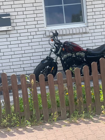 The image shows a red motorcycle parked on a paved area next to a white brick building in Großbeeren, Germany. The building has a dark mailbox with "Jarosch" and the number "21" on it, and a window with a grid pattern. A low wooden fence with vertical slats separates the paved area from a patch of grass and plants. The motorcycle is a Harley-Davidson Sportster, recognizable by its engine shape, fuel tank, and handlebars. The motorcycle's front wheel is turned slightly to the left, and its kickstand is down. The sun is shining, casting shadows on the ground from the fence posts. There are no people visible in the image.
