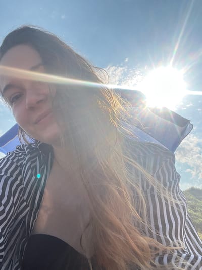 A young woman with long, light brown hair is pictured from the chest up, outdoors against a bright blue sky with scattered clouds and a harsh sun flare. She is wearing a black top and an open black and white striped shirt. She is looking towards the camera with a slight smile. The background suggests a hilly, vegetated area. The lighting indicates it is daytime, likely sunny.