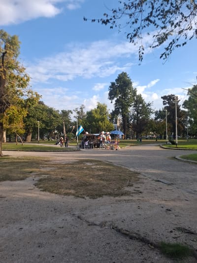 The image depicts a park in Villa Domínico, Argentina. A dirt or gravel path runs along the right side of the frame. To the left of the path, there is a grassy area with trees. In the middle ground, a group of people are gathered near a booth or vendor stand, with an Argentine flag visible. A blue sky with scattered clouds forms the backdrop.