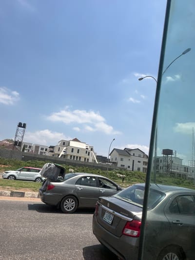 The image shows a view of Aso, Nigeria, from inside a vehicle, likely a car, looking out through a window. The scene features a clear blue sky with scattered clouds. In the middle ground, there are modern multi-story residential buildings. A utility tower with water tanks is also visible. On the right side of the image, the rear ends of several vehicles are positioned on a paved road. The gray car in the immediate foreground has a license plate visible. The perspective is tilted, adding to the dynamic nature of the shot.