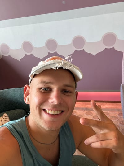 A man with a shaved head smiles for a selfie. He is wearing a blue tank top and a white cap with a fried egg design on it. He has a white wireless earbud in his left ear and is making a peace sign with his right hand. The background is a purple wall with a decorative pattern and a pink light strip. The image was taken in Huangpu District, China.