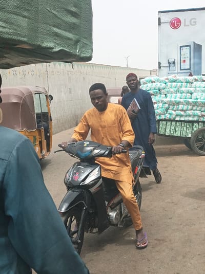 The image displays a daytime street scene in Fagge C, Nigeria. In the central foreground, a male of dark complexion, wearing an orange long-sleeved traditional tunic and matching trousers, is seated on a black and grey underbone motorcycle. He grips the handlebars, and his left foot, wearing a purple flip-flop, rests on the unpaved ground. A dark-colored watch is visible on his left wrist.
To the right and slightly behind the motorcyclist, another dark-complexioned male, dressed in a dark blue traditional garment and a reddish-brown cap, is walking towards the viewer's right. He holds a patterned book or folder in his left hand. The extreme foreground left shows the back and right shoulder of an individual wearing a dark teal long-sleeved garment.
To the left of the central motorcyclist, a brown three-wheeled auto-rickshaw (Keke Napep) is visible. A large object, partially visible in the upper left, is covered with a green tarp. In the background, a light grey concrete wall, adorned with decorative patterns featuring small triangles and star-like shapes, spans the mid-ground. To the far right, a white truck bearing an "LG" logo and a refrigerator graphic is present. Behind this truck, numerous white sacks or bags are stacked high on a two-wheeled cart or trailer. The ground is an unpaved, light brown surface, and the sky is bright and hazy.