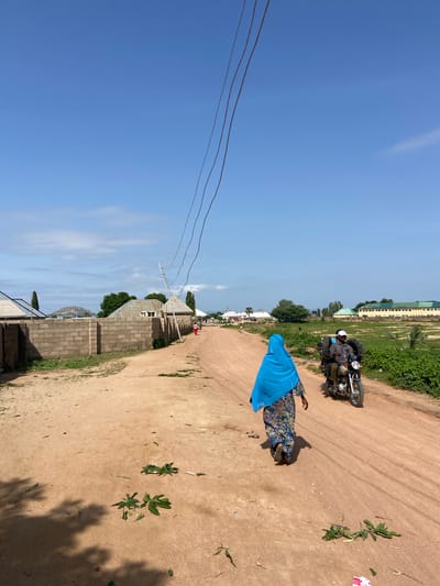 In Kuson, Nigeria, a woman wearing a blue hijab and a colorful patterned dress walks away from the camera on a dusty dirt road. She is followed closely by a man riding a motorcycle, who is carrying two spare tires on the back. The road is lined with a low brick wall on the left, behind which are houses with corrugated metal roofs. Trees and a hill are visible in the distance. On the right side of the road, there is a large field with green vegetation and a long, light-colored building with a green roof. Power lines stretch across the clear blue sky. Scattered leaves and debris are visible on the road.