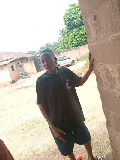 The scene is set in a semi-urban or rural residential area within Eziudo, Nigeria. A young individual, appearing male, with short dark hair and dark skin, stands in the immediate foreground, slightly angled to the left, with their head turned towards the viewer. They are wearing a black short-sleeved t-shirt with a red and blue outlined "C" logo on the chest, dark blue shorts, and a black and white striped slide sandal on their visible left foot. The individual's right hand rests on a rough-textured, light brown concrete pillar or wall that occupies the right side of the frame. A red plastic cup is on the ground near their left foot.
The ground is unpaved, consisting of light brown dirt and sparse dry vegetation. In the mid-ground, a light-colored (possibly silver) minivan or SUV is parked. Behind it, a low, light brown concrete wall extends horizontally. Further left, a single-story building with a light brown facade and a darker roof is visible, along with a closed dark metal gate and a light blue or grey motorcycle parked nearby. Dense green foliage and tall trees, including a prominent leafy tree, are present in the background, under a bright, likely overcast sky.