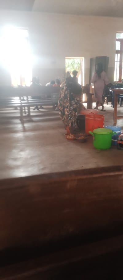 The image depicts an indoor scene that appears to be a community gathering or meeting space in Maigizo, Nigeria. In the foreground, a concrete floor is visible, with a dark wooden surface in the lower portion of the frame. To the right, there are several plastic containers of various colors, including a bright green one with a lid, a larger orange one with a lid, and a blue cooler. Small bowls containing what looks like food are placed near these containers.
In the midground, a woman in a patterned dress is standing near the containers. Behind her, a group of people are seated on long wooden benches, facing towards the left side of the image. Some individuals are obscured by the benches and the lighting.
The background features a large window on the left side, which is heavily overexposed with bright sunlight streaming through. Another doorway or opening is visible in the center, with foliage suggesting an outdoor view. On the right side of the background, there is a tall, dark cabinet or structure, and a wooden table with chairs is partially visible. Another person, dressed in light-colored clothing, is standing and walking towards the right. The overall lighting is dim, with strong light from the window creating shadows and reducing visibility in some areas. The setting suggests a casual, communal environment.